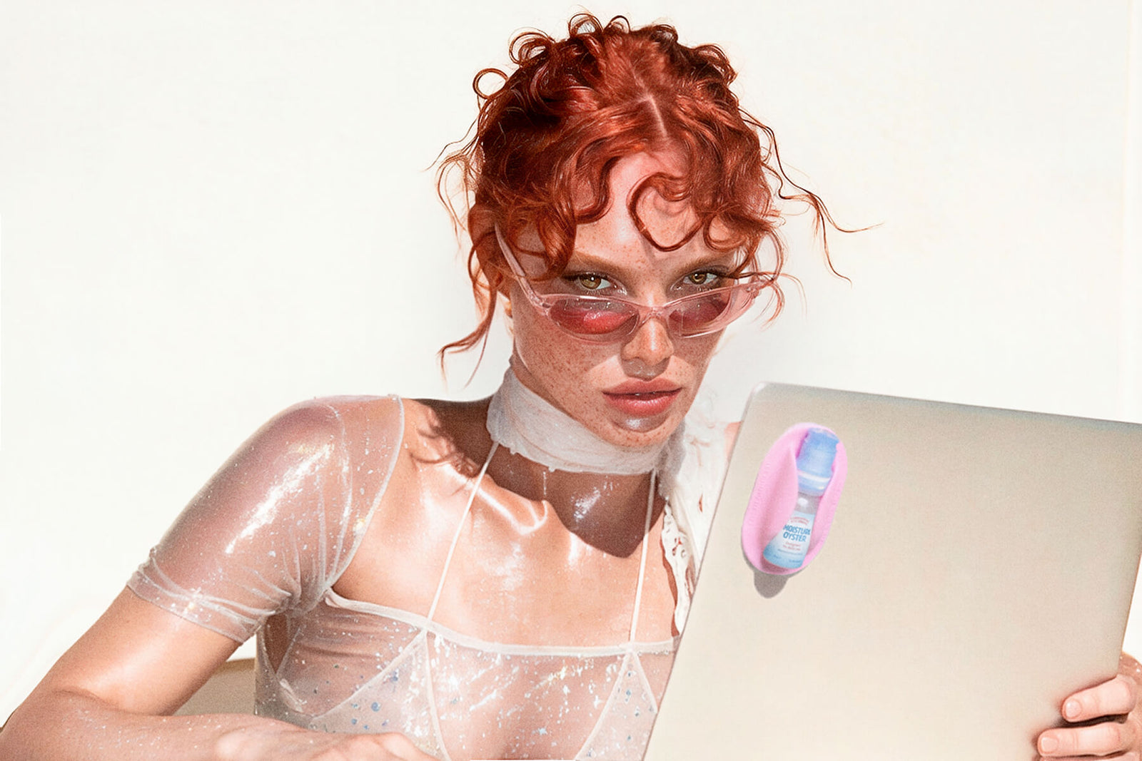 A model with vibrant red hair and pink-tinted sunglasses wears a sheer, wet, sparkly white top and a white choker, holding a silver laptop with the Moisture Oyster eye drops attached to the screen.