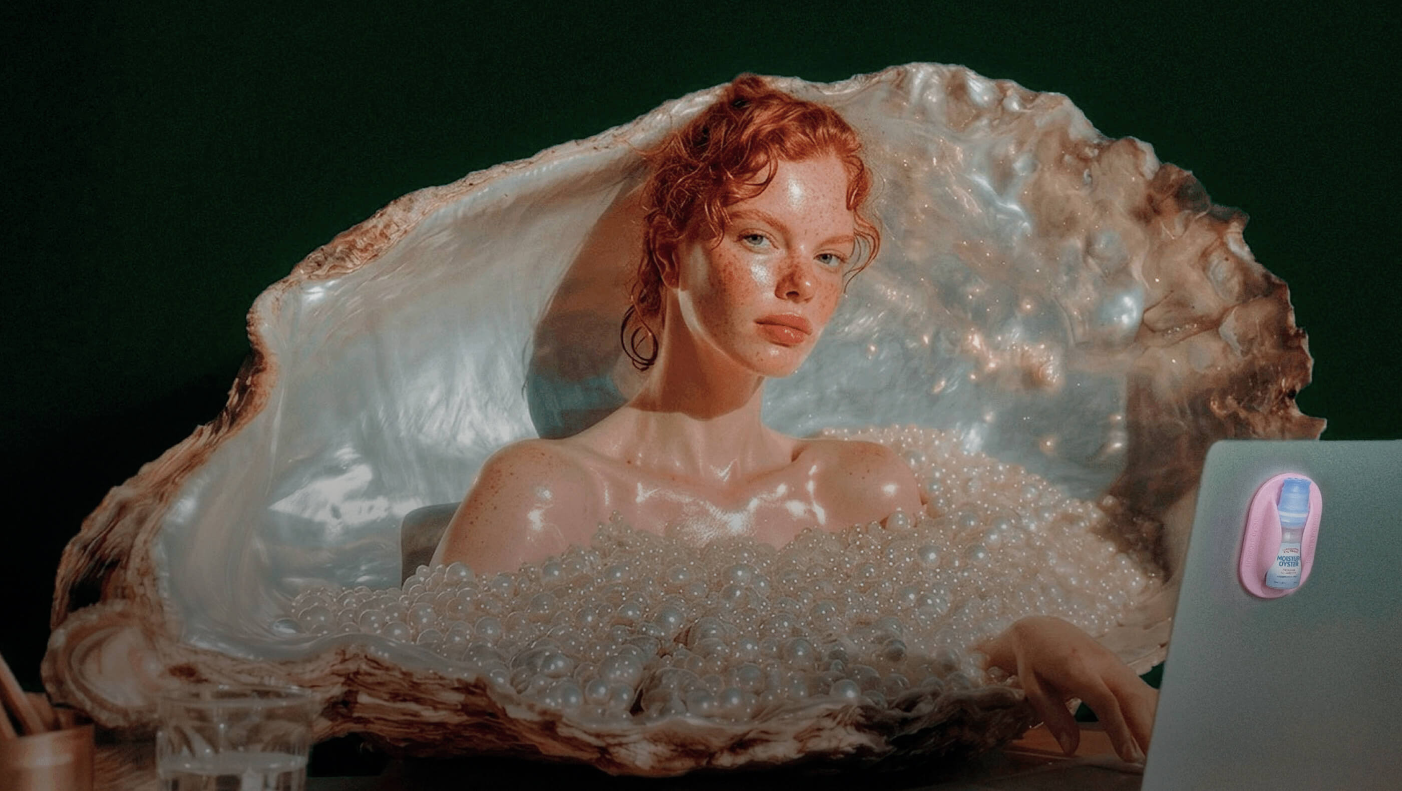 A large realistic shell sculpture containing a painted female figure with red hair, set against a dark background with a laptop partially visible on the right.