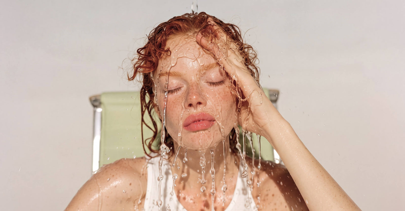 A woman with wet red hair and closed eyes under running water, wearing a white tank top and sitting in a light green chair against a plain light background.